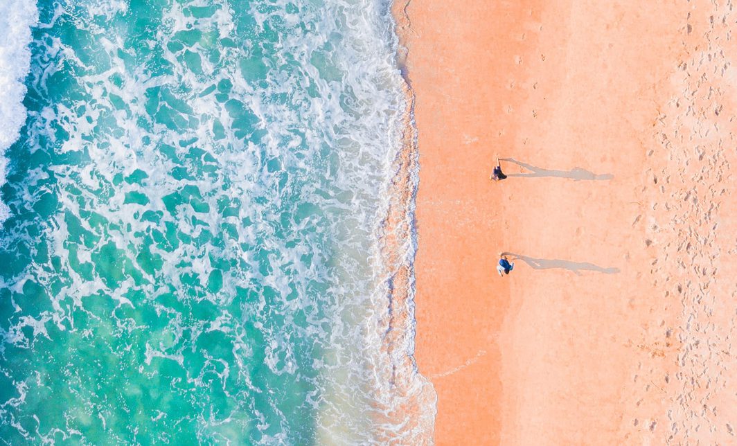Turquoise waters washing up on a yellow sand beach with two people walking the coastline.